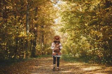 Poster Stylish woman hipster with backpack in hat walking in sunny autumn woods. Young female traveler hiking in fall forest, beautiful moment. Travel and wanderlust concept, space for text. Back view © sonyachny