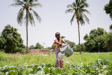 Pictoresque image of a cute little African girl watering vegetable plants in a field surrounded by...