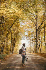 Obraz premium Stylish woman traveller in hat with backpack walking with cute dog on road in sunny autumn woods. Young female hipster hiking with swiss shepherd white dog. Travel and wanderlust concept