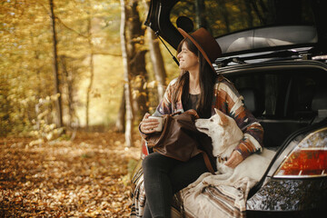 Stylish woman traveller with backpack sitting with cute dog in car trunk in sunny autumn woods....