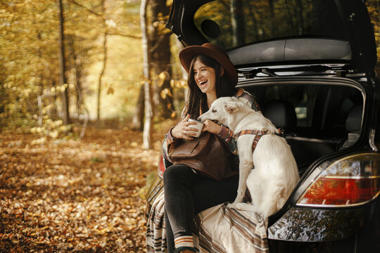 Stylish woman traveller with backpack sitting with cute dog in car trunk in sunny autumn woods. Young hipster female traveling with swiss shepherd white dog. Travel and road trip with pet