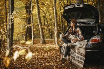 Stylish woman traveller with backpack sitting with cute dog in car trunk in sunny autumn woods. Young hipster female traveling with swiss shepherd white dog. Travel and road trip with pet