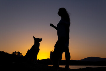 Silhouette of woman playing with a dog at sunset in front of a lake. Love for animals. Selective focus. Copy space.