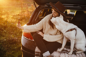 Autumn road trip with pet. Stylish hipster woman taking selfie photo with cute dog in car trunk in warm sunset light. Young female in hat and sweater using phone and travelling with sweet white dog