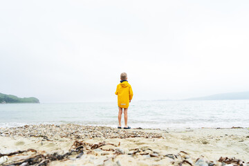 A little boy child in a yellow windbreaker stands on the sandy shore of the ocean all alone. © Aboltin