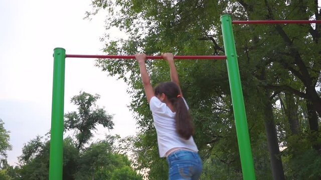 A teenage girl is engaged on horizontal bars. Playground. The child goes in for sports. City block. Leisure park. Workout for health. Competition.
