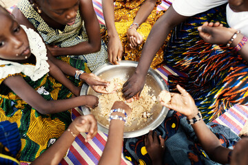 Group of black African girls sitting on a mat, dividing their frugal meal, eating with their hands from a large metal plate - obrazy, fototapety, plakaty
