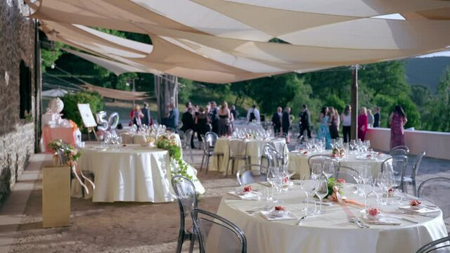 Blurred wedding guests waiting for married couple on foreground of served festive tables decor. Beautiful round tables decorated with flowers bouquets and elegant tableware and cutlery. Wedding party