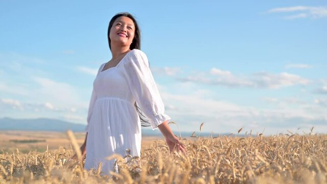 Young Woman in white dress standing on golden wheat field at sunny day, touching gently the wheat spikes. High quality 4k footage