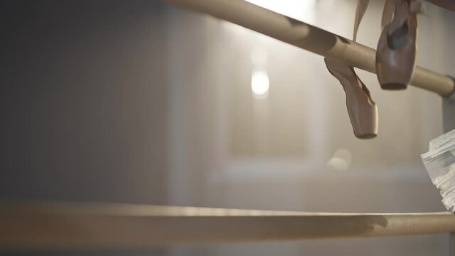 Beige pointe shoes hanging on ballet barre in backlit fog as unrecognizable ballerina taking pointes leaving. Ballet dancer on rehearsal in ballroom studio indoors. Slow motion