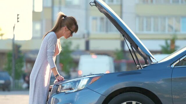 Young woman driver opening car bonnet inspecting broken motor on a city street. Vehicle malfunction concept.