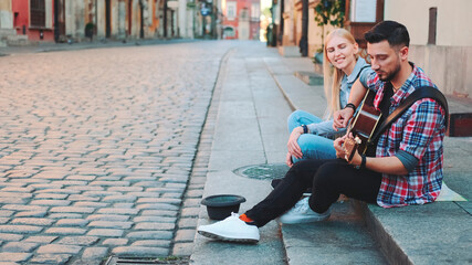Young couple of street singers in old part of the city sitting on sidewalk. Man playing guitar and woman singing.