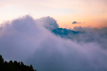 Naklejka na ściany i meble Dark Caucasus mountains peak shrouded in fog and fluffy clouds at colorful sunset. Pink and yellow cloudy skyscape. Stunning tranquil scene in Europe. Beauty in peaceful nature.