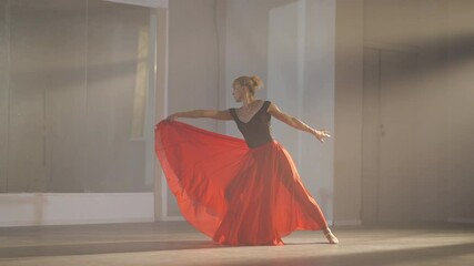 Slim woman in long scarlet red skirt and pointes jumping and bending in slow motion in backlit fog. Wide shot of graceful elegant female ballet dancer rehearsing dance in studio indoors