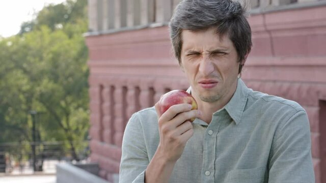 Young adult male shopper customer wants to eat delicious juicy ripe apple, sniffs, makes grimace of disgust. Fruit has bad smell, is spoiled, rotten, nitrates used, unsuitable for eating. Unfair trade