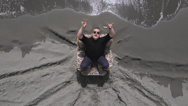 Man model rapper in sunglasses poses in armchair ON sandy beach with trace decorations and white rolling sea waves aerial view. Montenegro, Ulcinj, Velika Plaza
