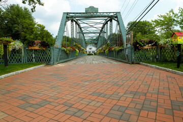 A red brick road leads to an iron bridge with wooden planks.  Hanging plants and flowers.