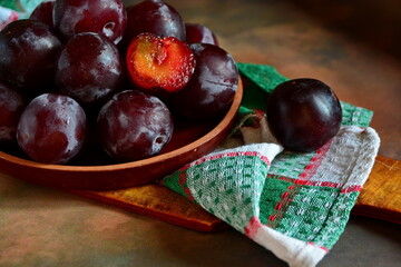 Black Cherry Plums and one slice in bowl and one pieces nearby close up on checkered towel on cutting on brown dark background. Side view