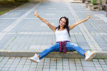 Fototapeta na wymiar Portrait of young beautiful brunette woman with long hair in blue jeans and white t-shirt sitting on sidewalk on city street. Lifestyle cheerfully spread her arms to the sides. Happy caucasian woman