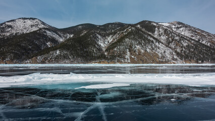 Cracks, snow spots, hummocks are visible on the blue ice of the frozen lake. The wooded slopes of the coastal mountains are covered with snow . Reflection on a smooth surface. Winter day. Baikal