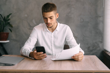 a young male office worker examines the documents and makes a call or sends a message to the client by phone. A guy in a white shirt sitting at a desk with a smartphone.
