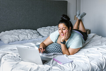 Fototapeta premium Curvy latin woman lying on bed using computer in Latin America, plus size female
