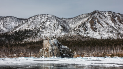 Fototapeta premium A granite rock with bizarre outlines and a flat top rises above a frozen lake. There is snow at the base, hummocks. Reflection on the ice. The background is a snow-covered mountain range. Baikal