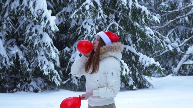 Young girl wearing santa claus hat and inflate red balloons. Winter forest in snow. woman jump, laugh. joy on happy Christmas eve. New Year's fun. pretty girl in red hat. copy space for advertising.