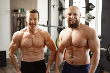 Fototapeta na wymiar Two shirtless men standing in gym. Young man and his senior father exercise together in gym
