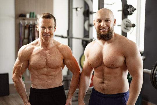 Two shirtless men standing in gym. Young man and his senior father exercise together in gym