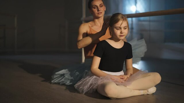 Beautiful Caucasian girl in tutu smiling sitting on floor in ballet studio as young woman adjusting hairdo at background talking. Happy student and teacher in dancing studio indoors. Childhood and art
