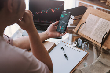 Young investor learning to trade the stock market and the cryptocurrency market at home ,Asia young trading making stock market analysis on smartphone with laptops featuring stock tickers or graphs. 