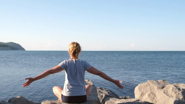Girl sits on the stones by the sea and practices yoga in the early morning. Young woman meditates while sitting in lotus position by the ocean at sunrise. Concept of nature, health, solo activity
