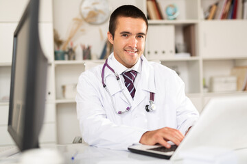 Positive man doctor sitting at workplace with computer in her office