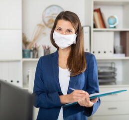 Fototapeta na wymiar Smiling woman in protective medical mask working with papers and laptop in office
