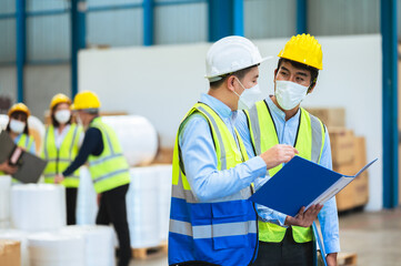 Team engineers and foreman wear a mask, hard hat, and vest. Standing, consult discuss industrial production management through data files with in mask factory. Employees working in the background.