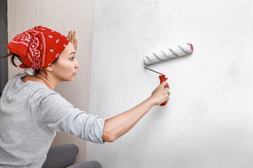 Asian woman applying primer and glue with a wide roller on the plastered surface of the wall for...