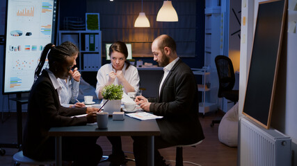 Diverse multiethnic business teamwork overworking in office meeting room analyzing financial graphs paperwork late at night. Coworkers brainstorming project ideas solving company problem