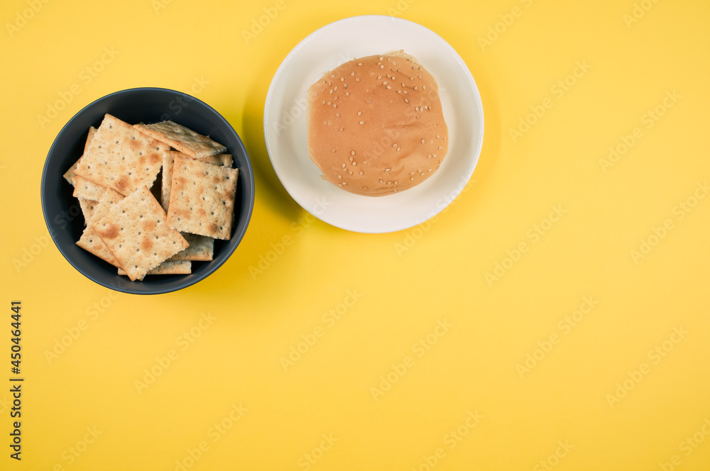 Poster Bowl of square crackers and bread bun on a plate isolated on yellow background