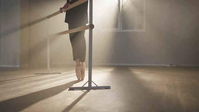 Unrecognizable slim ballerina in black suit and pointes walking to barre in backlit fog and standing in a la quatrieme devant. position Talented graceful Caucasian woman rehearsing steps in studio
