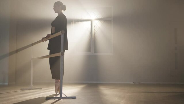 Wide shot of confident slim ballerina in pointes walking on tiptoes to barre in backlit fog. Gorgeous Caucasian brunette woman in black suit rehearsing in smoke dance studio