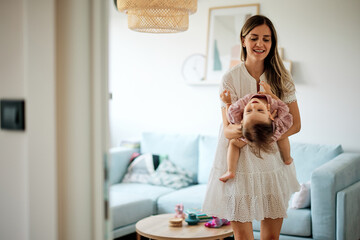 A cheerful mother standing in the living room with her daughter in her hands and playing with her.