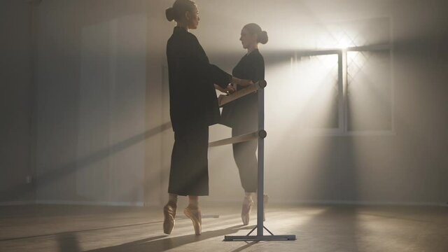Two graceful slim ballerinas walking to barre on tiptoes and bending back in croise derriere. Confident Caucasian young women rehearsing dance in backlit fog in dancing studio. Femininity and art