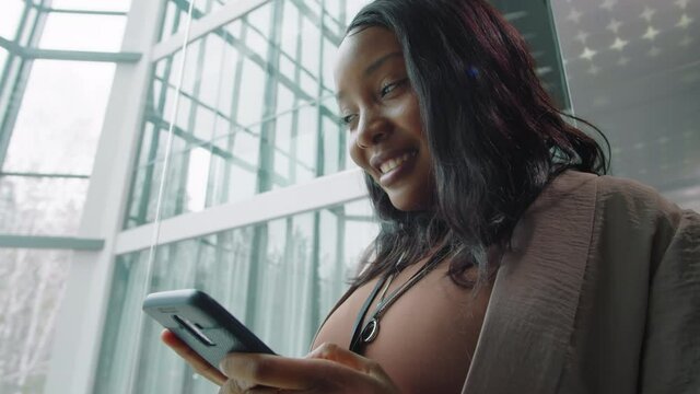Cheerful Afro-American businesswoman smiling and typing on smartphone while riding elevator in office center