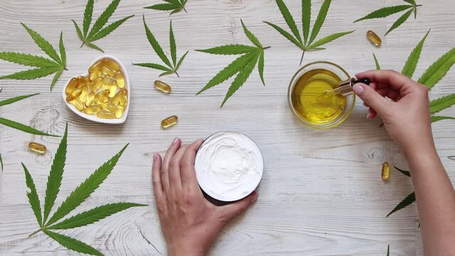 Woman dripping CBD oil into cream at light wooden table surrounded by cannabis leaves. Top view.