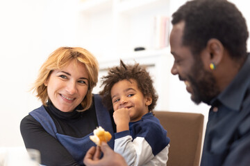 small child, half African and half Caucasian, in his mother's arms, looking at his father with a playful expression, moments of joyful life of a mixed race family