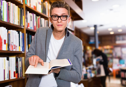 Surprised cheerful positive glad male student standing in library, pointing at something in book