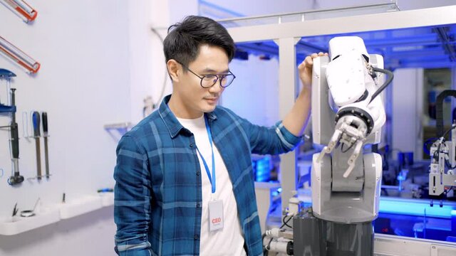 Young Asian male Industry CEO in glasses standing touching an automation robotic arm machine that are moving while testing for work, then looking at camera with arms folded and smiling in a workshop.