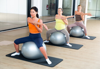 Women maintaining active lifestyle exercising with gym ball during group pilates class in modern...