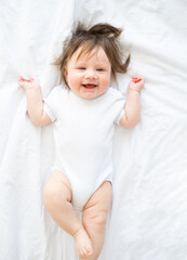 funny baby girl in white bodysuit smiling and lying on a white bedding at home. top view.